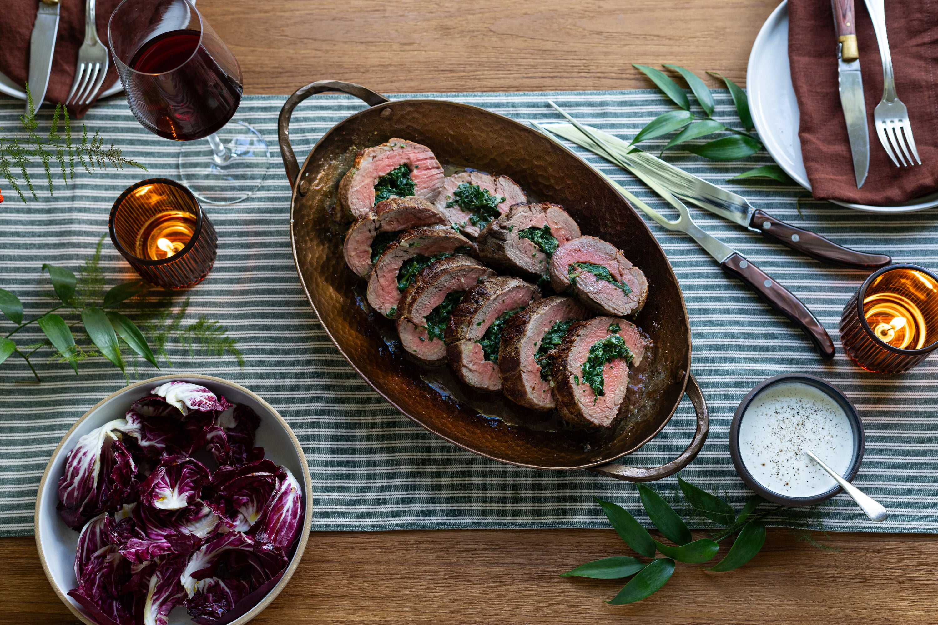 Roasted beef medallions with greens, surrounded by red wine, salad, and candles on a striped tablecloth.